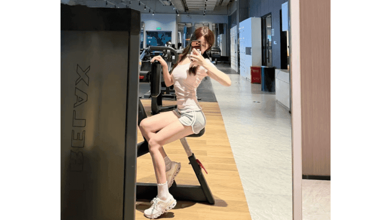 Stripchat youmibaby11 taking a mirror selfie at a sports gym wearing a T-shirt and short pants to showcase her fit body and beautiful legs